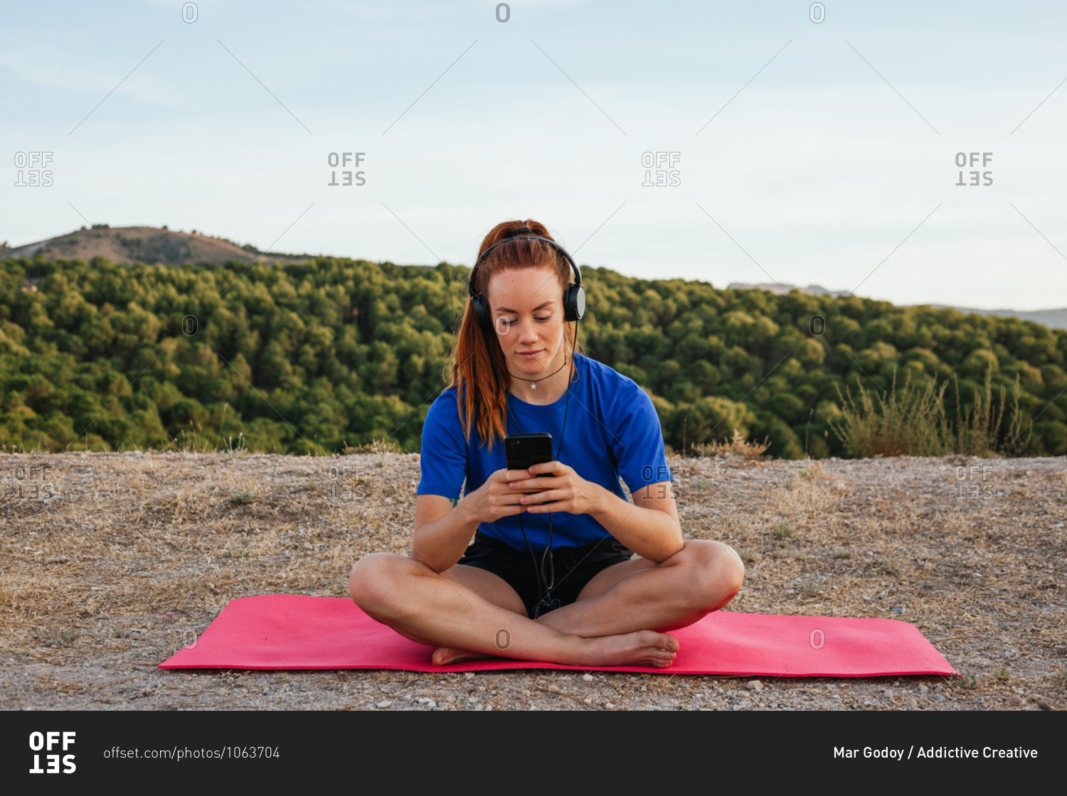 Serene female in sportswear sitting on mat and choosing songs from playlist while enjoying calm music in headphones after doing yoga in nature