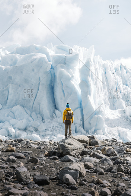Back view of unrecognizable tourist in outerwear with backpack standing on stony terrain against gigantic blue ice formation while visiting Perito Moreno glacier in Argentina