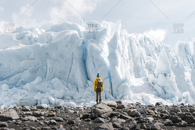 Back view of unrecognizable tourist in outerwear with backpack standing on stony terrain against gigantic blue ice formation while visiting Perito Moreno glacier in Argentina