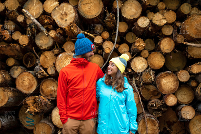 Happy young man and woman in warm outerwear and hats embracing and looking at each other while standing near heap of cut tree logs in forest