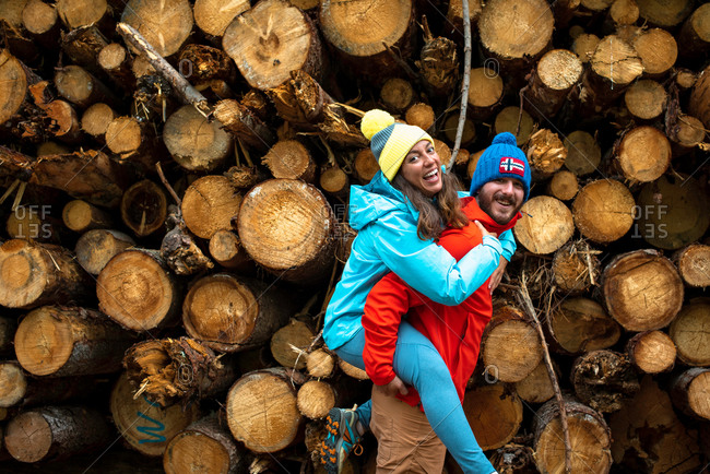 Happy young bearded man in warm clothes carrying cheerful girlfriend while spending time together in forest near pile of logs