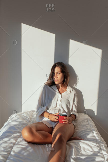 Happy young lady in white shirt sitting on comfortable bed and smiling while drinking cup of coffee in sunny morning looking away