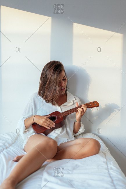 Alluring young female musician in white shirt sitting on comfortable bed and playing ukulele in sunny morning