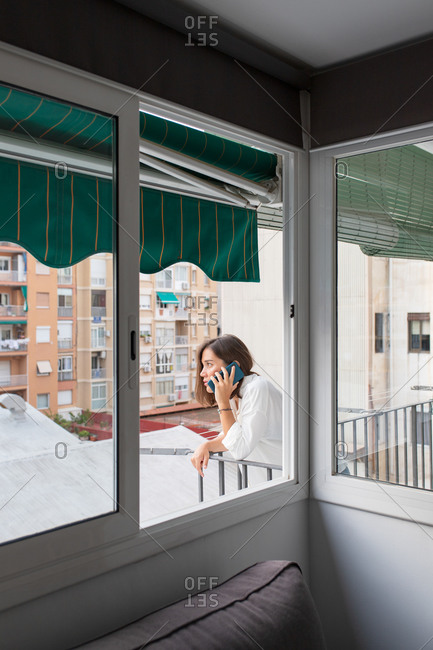 Side view of young woman in casual outfit leaning on balcony railing and having phone conversation in daytime