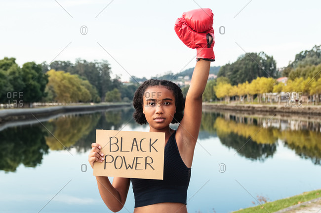 Determined young African American sportswoman in boxing gloves demonstrating placard with Black Power inscription after workout in park near lake