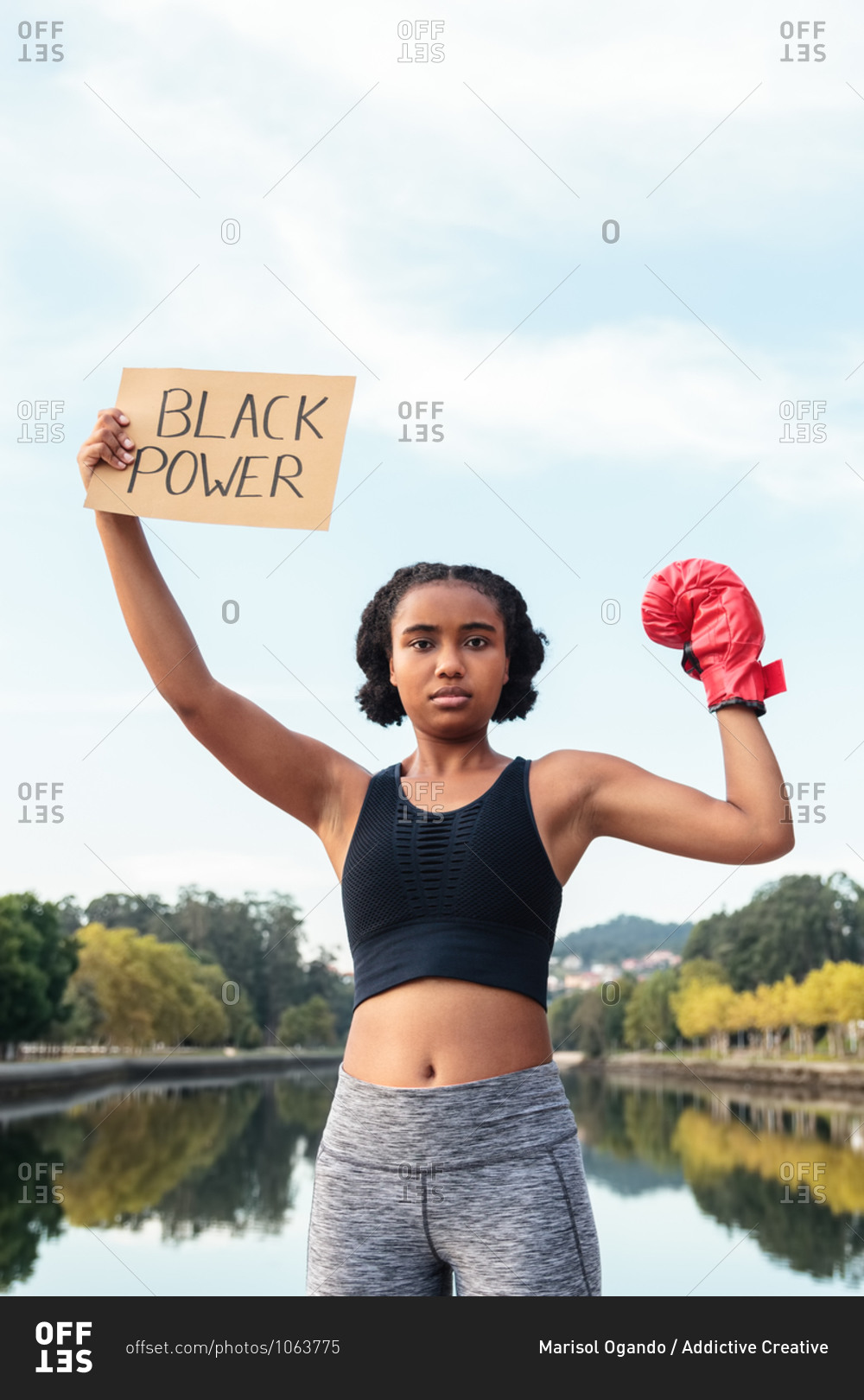 Determined young African American sportswoman in boxing gloves demonstrating placard with Black Power inscription after workout in park near lake