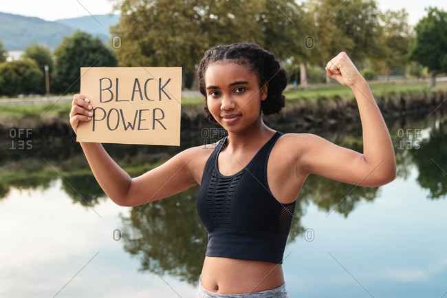 Cheerful young sporty ethnic lady in activewear demonstrating biceps and placard with Black Power text looking at camera after outdoor training near pond in park
