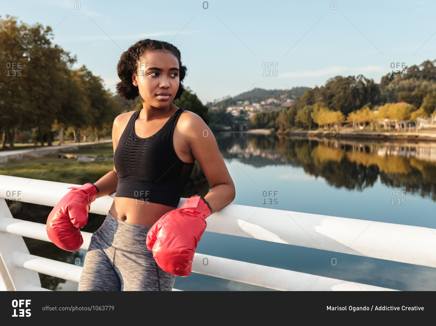 Contemplative young ethnic sportswoman in boxing gloves and sports clothes looking away while leaning on fence above river reflecting trees