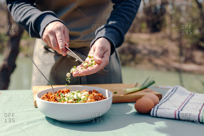 Unrecognizable male cook in apron standing at table in yard in countryside and adding green onions in bowl with appetizing minced meat