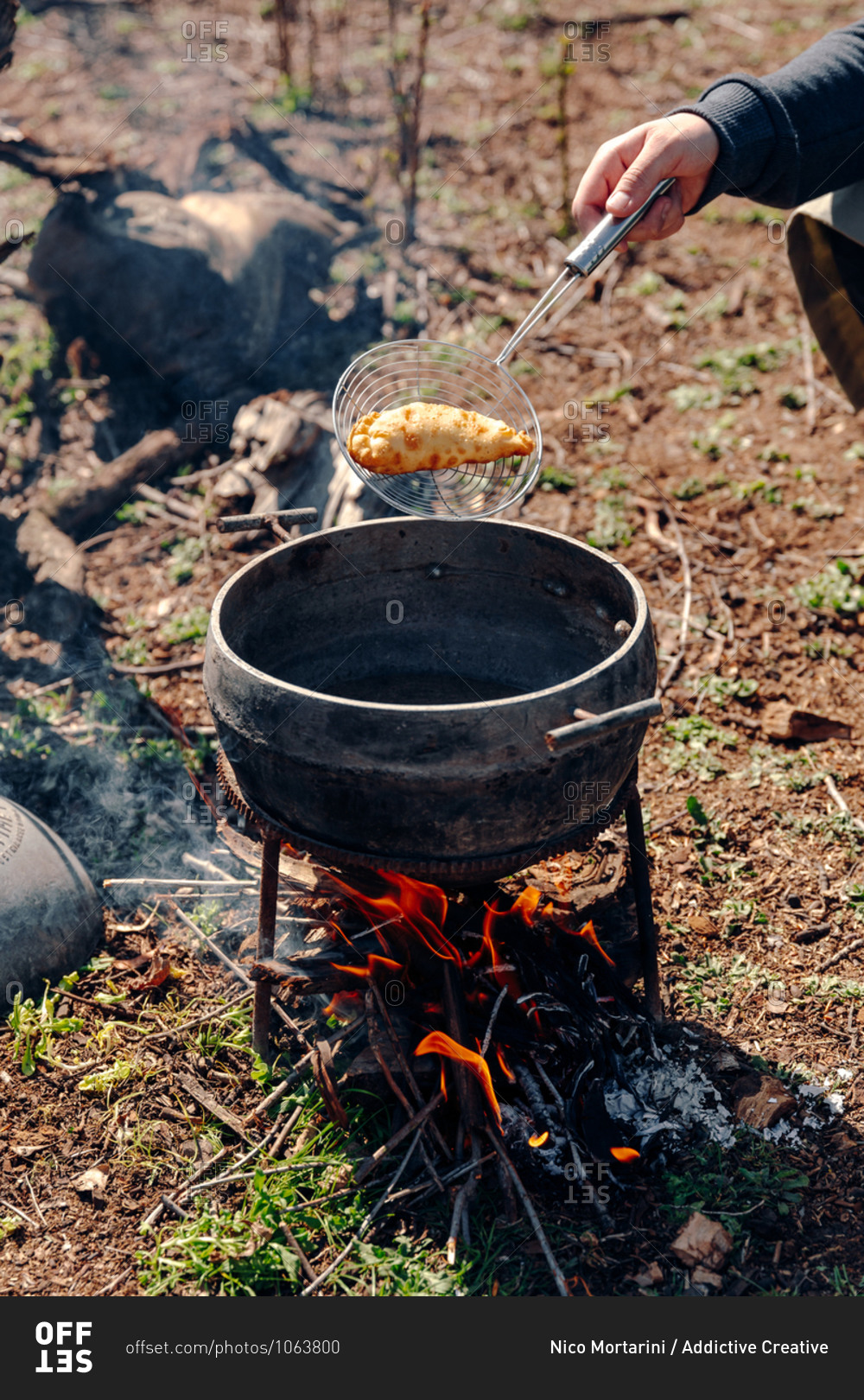 Unrecognizable cook preparing delicious pasty in boiling oil in pot on camping fire in countryside