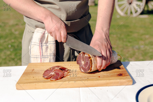 Unrecognizable crop cook standing at table in countryside and cutting delicious bresaola on wooden cutting board