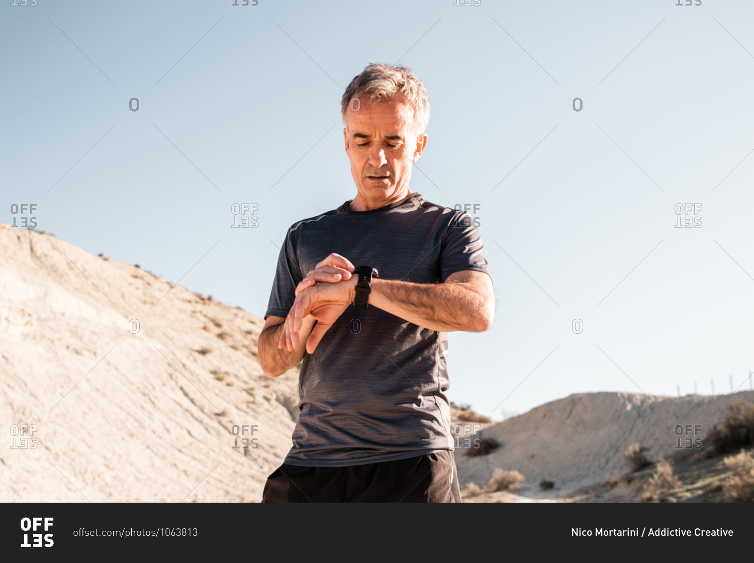 Concentrated middle aged male athlete in activewear checking time on smartwatch while standing in hilly terrain after workout