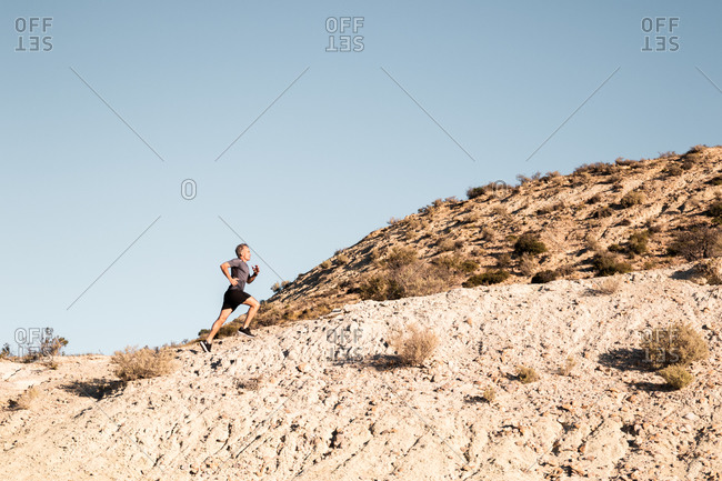 Side view of active male jogger running uphill in sandy semi desert terrain during workout in sunny morning