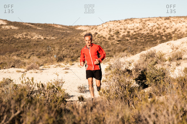 Front view of active male jogger running uphill in sandy semi desert terrain during workout in sunny morning