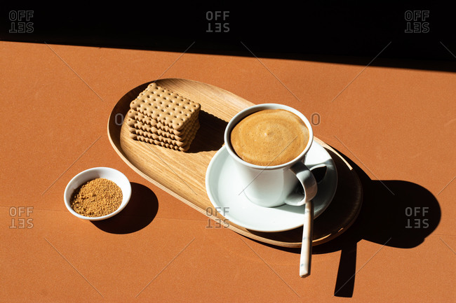 From above of ceramic cup of tasty coffee near crunchy crackers on wooden plate near bowl with cane sugar in cafe in sunlight