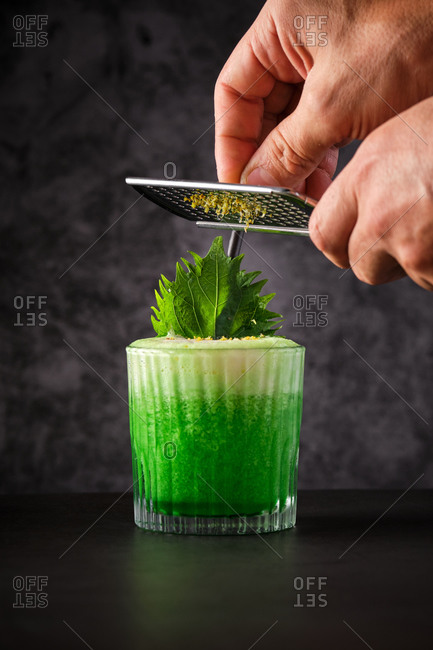 Crop unrecognizable bartender grating citrus zest on green cocktail decorated with fresh mint leaves and served on black table