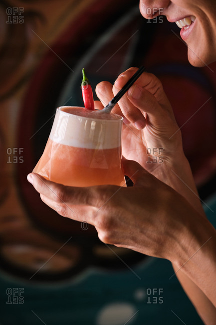 Crop anonymous bartender holding glass of fresh foamy drink decorated with straw and chili pepper