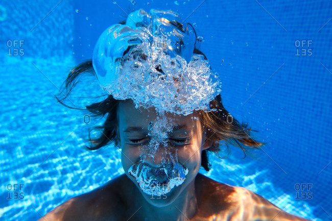 Underwater shot of cheerful teenage boy diving into blue swimming pool water with eyes closed and smiling happily