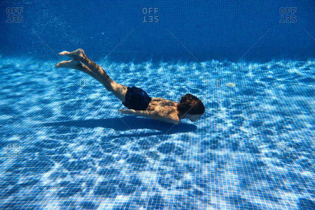 Underwater shot full body energetic teenage boy in swimming trunks diving into clean pool water in resort