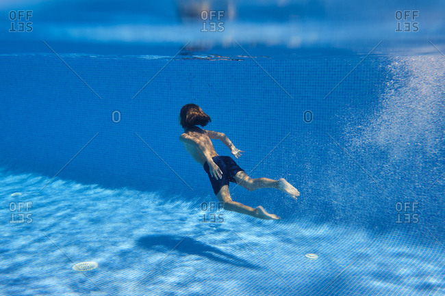 Underwater shot full body energetic teenage boy in swimming trunks diving into clean pool water in resort
