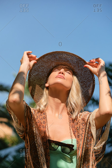 Serious young blond haired slim female tourist touching hat with eyes closed while lounging in sunlight under blue cloudless sky