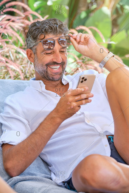 Cheerful young adult bearded male in summer clothes watching funny video on smartphone while relaxing on sunbed in tropical garden