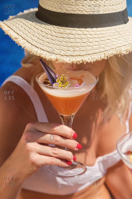 From above of crop anonymous female traveler in straw hat and white bikini enjoying exotic fruit cocktail decorated with flowers while relaxing on poolside during summer holidays