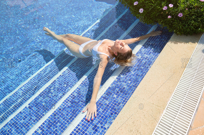 From above of happy slim female traveler in swimwear lying in swimming pool and sunbathing with closed eyes