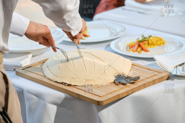 Crop anonymous waiter cutting crust of fish baked in salt dough while serving food to clients in modern restaurant