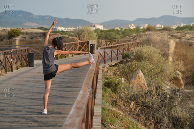 Back view of sporty female in activewear stretching while standing on wooden path