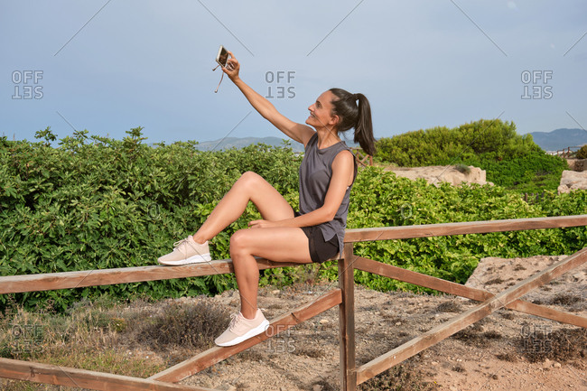 Side view of cheerful happy sporty female using mobile resting on wooden fence near green plants