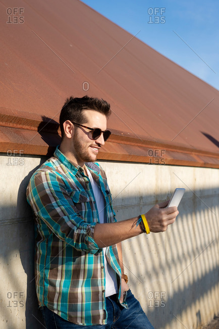Positive young male in checkered shirt and sunglasses taking selfie on modern mobile phone while standing near shabby concrete wall on street