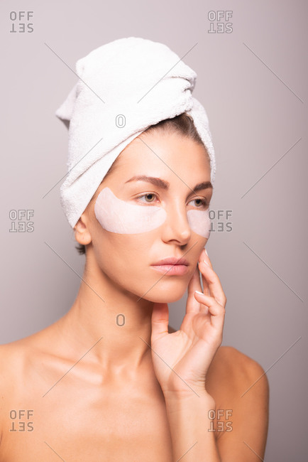 Charming female model with towel turban on head and hydrating eye patches on face standing on violet background in studio and looking away