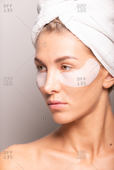Charming female model with towel turban on head and hydrating eye patches on face standing on violet background in studio and looking away
