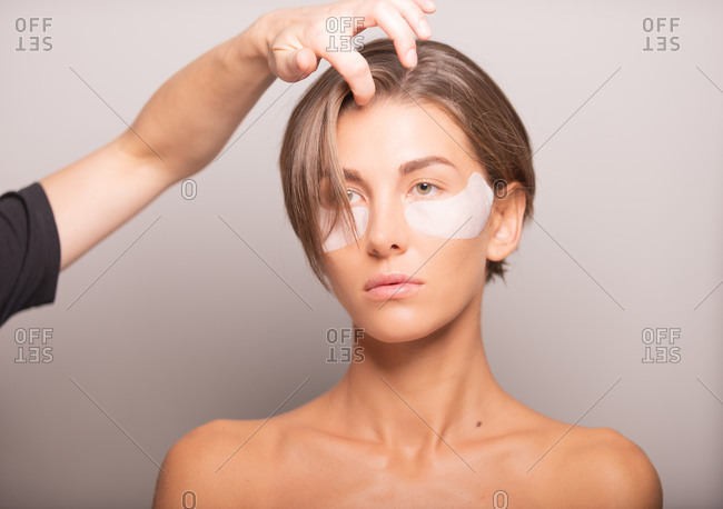 Cropped unrecognizable female hairstylist adjusting short hair of model standing with eye patches on violet background in studio