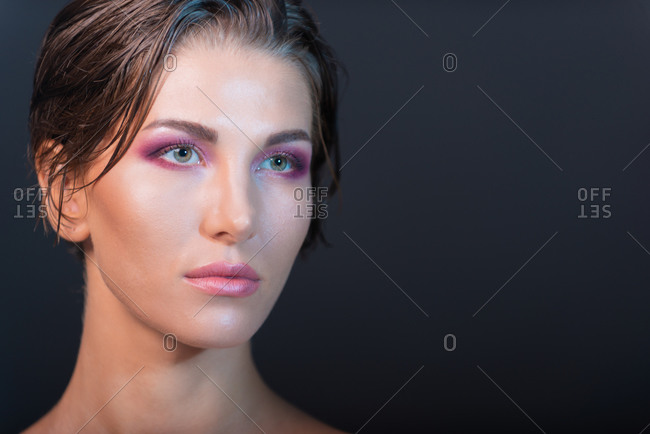 Charming female model with wet short hair and bare shoulders standing on violet background in studio and looking away