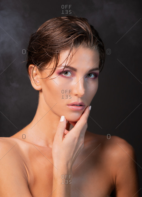 Charming female model with wet short hair and bare shoulders standing on violet background in studio and looking at camera