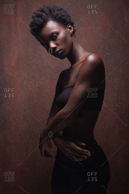 Sensual African American female wearing tight black top with bare shoulders standing with arms crossed in dark studio and looking at camera provocatively