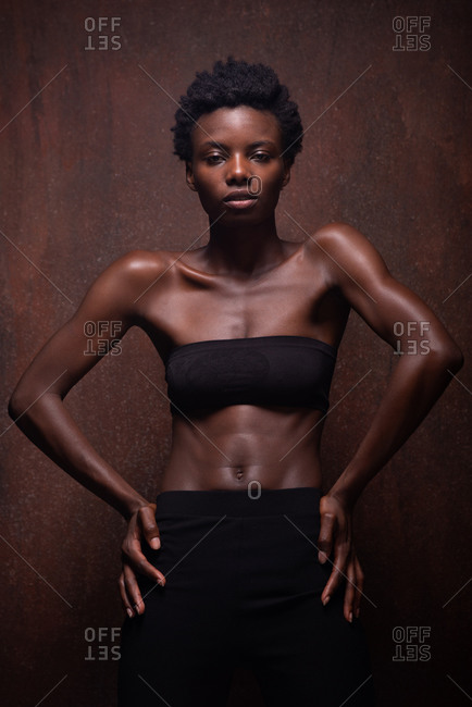 Seductive African American female model wearing black top with bare shoulders with open arms in front and looking at camera during photo session in dark studio