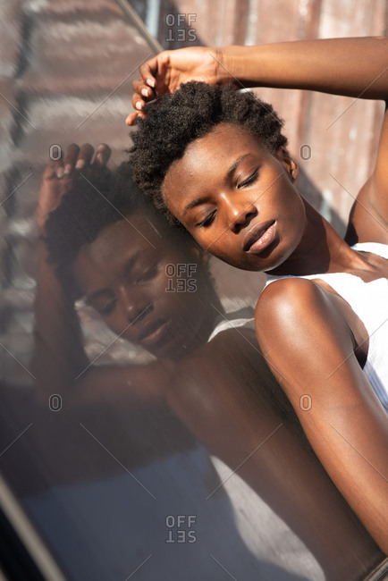 Side view graceful African American female model in white shirt laying back with eyes closed on construction on sunny roof and touching head gently