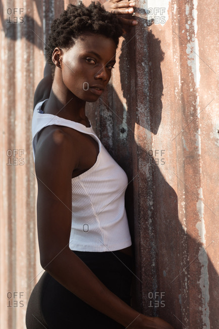 Attractive slim African American female in white shirt raising arm while leaning on grunge wall and looking at camera calmly