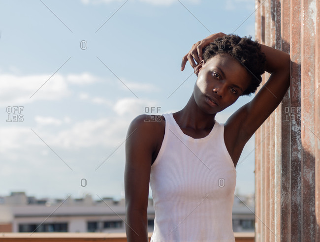 Attractive slim African American female in white shirt raising arm and touching head while leaning on grunge wall and looking at camera calmly