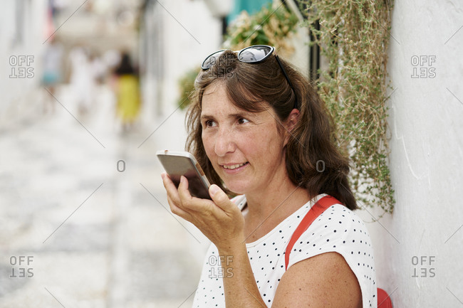 Content adult female in casual summer wear recording audio message on modern mobile phone while standing on sunny pedestrian street