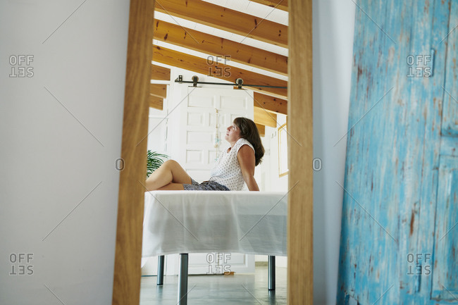 Side view serene adult female in casual summer wear resting with legs crossed on table on sunny terrace and reflecting in mirror