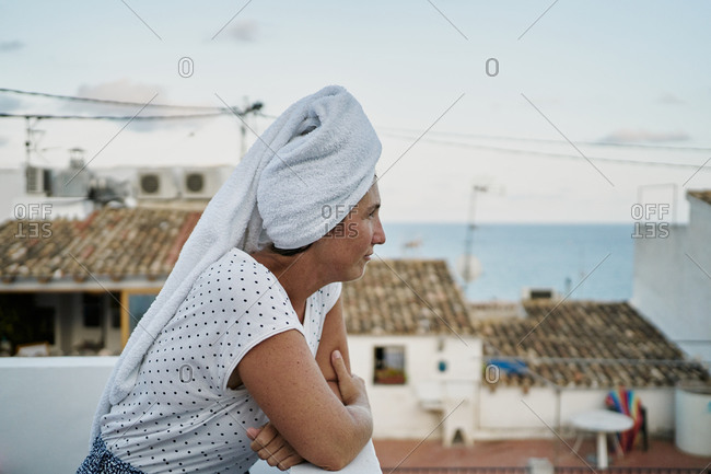 Side view calm adult female in casual shirt with bath towel on head leaning on balcony railing and enjoying white town views