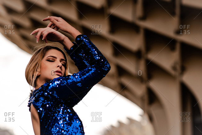 Low angle of young reflective trendy woman with makeup in blue apparel standing with raised arms and closed eyes under building roof