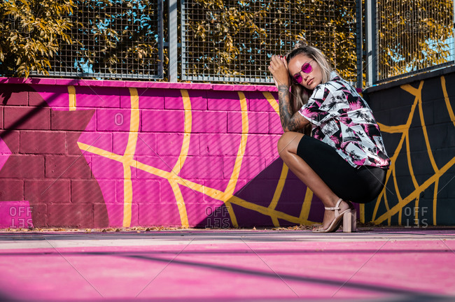 Ground level side view of young stylish female in ornamental clothes squatting on bright sports ground near grid fence and looking at camera
