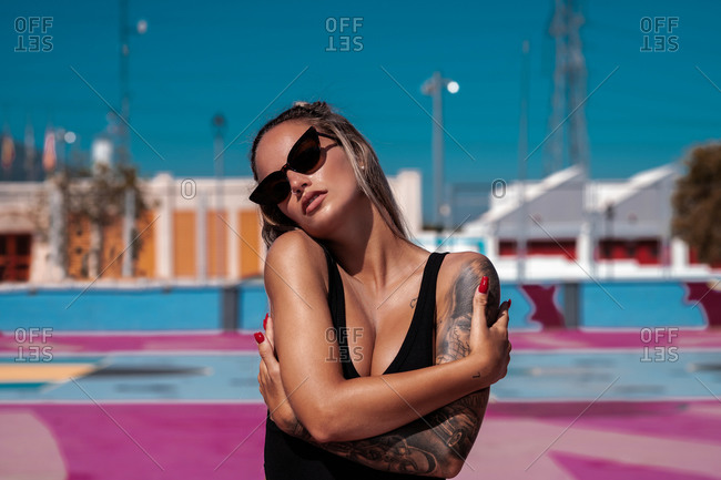 Young trendy female in modern sunglasses embracing herself on fenced pink sports ground in daylight