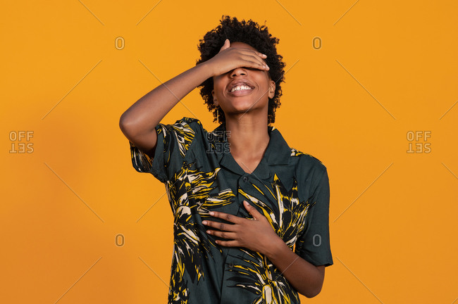 Young happy African American female in trendy wear with Afro hairstyle standing covering eyes standing on studio with orange background
