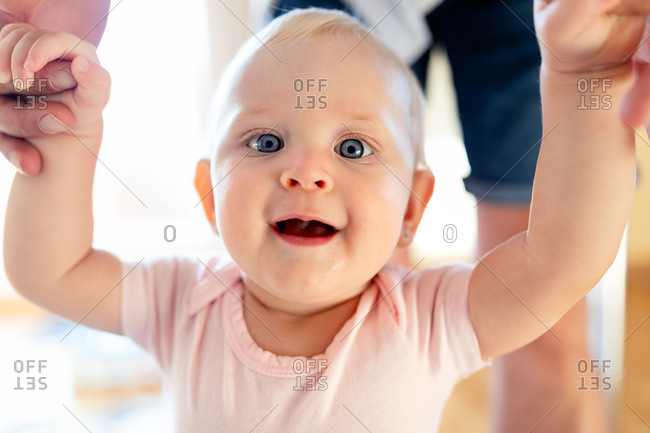 Cute baby with blond hair and light blue sky looking at camera with mouth opened while holding hands of faceless parent